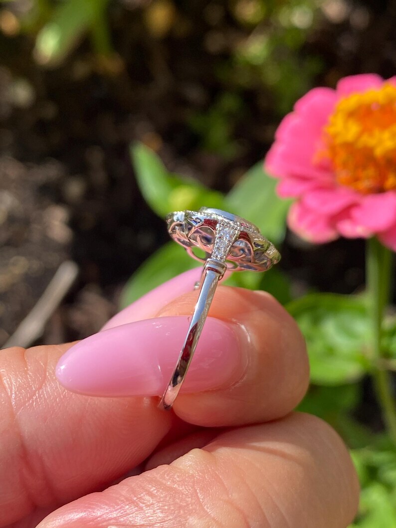 May include: A silver-toned ring featuring a central oval-shaped stone surrounded by a halo of smaller clear stones. The metal band displays intricate filigree detailing along the gallery and small stones set into the shoulders of the ring.