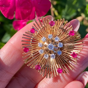 Puede incluir: Un broche de oro con un diseño de rayos de sol que presenta un grupo de diamantes en el centro y rubíes rosas alrededor del borde.
