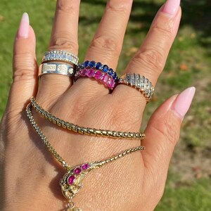 May include: A collection of gold jewelry, including a necklace with a heart-shaped pendant and rings with gemstones. The rings feature diamonds and sapphires. The necklace has a ruby-colored gemstone. The jewelry is displayed on a hand.