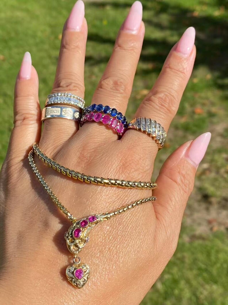 May include: A collection of gold jewelry, including a necklace with a heart-shaped pendant and rings with gemstones. The rings feature diamonds and sapphires. The necklace has a ruby-colored gemstone. The jewelry is displayed on a hand.