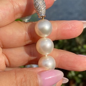 Pode incluir: Close-up de um colar de p&eacute;rolas com um fecho cravejado de diamantes. As p&eacute;rolas s&atilde;o grandes, redondas e de cor branco creme. O fecho &eacute; prateado e apresenta um design em espiral com pequenos diamantes brilhantes.