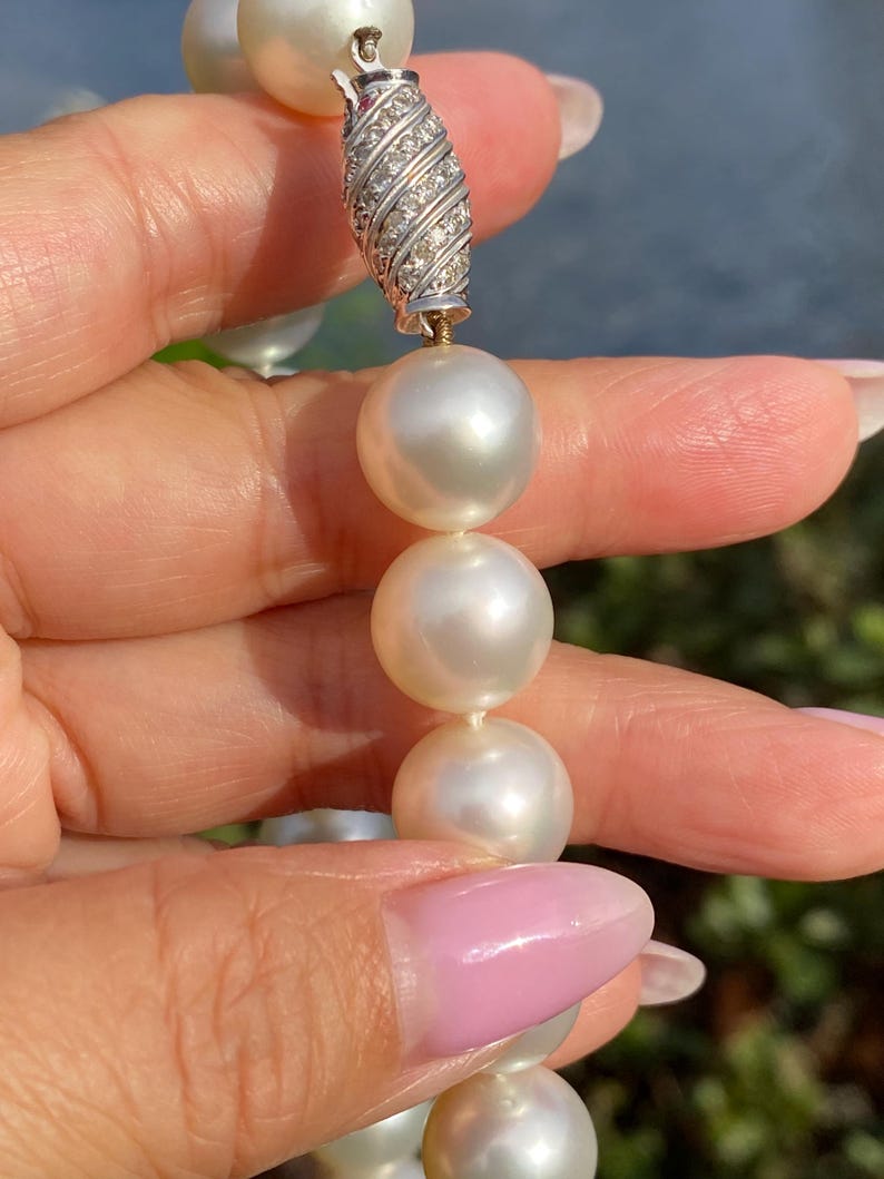 Pode incluir: Close-up de um colar de p&eacute;rolas com um fecho cravejado de diamantes. As p&eacute;rolas s&atilde;o grandes, redondas e de cor branco creme. O fecho &eacute; prateado e apresenta um design em espiral com pequenos diamantes brilhantes.