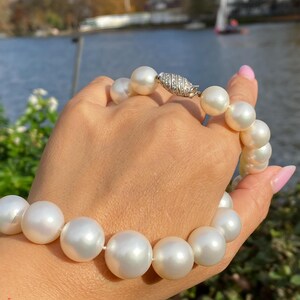 Pode incluir: Um colar de p&eacute;rolas com grandes p&eacute;rolas brancas redondas. O colar &eacute; preso com um fecho prateado adornado com pequenas pedras brilhantes. As p&eacute;rolas s&atilde;o enfiadas, criando uma pe&ccedil;a de joalheria cl&aacute;ssica e elegante.