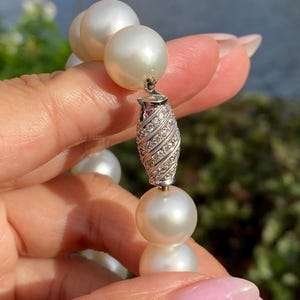 Pode incluir: Um close-up de um colar de p&eacute;rolas com um fecho decorativo. As p&eacute;rolas s&atilde;o grandes, redondas e brancas. O fecho &eacute; prateado e incrustado com pequenas pedras brilhantes. O fundo est&aacute; desfocado, com ind&iacute;cios de &aacute;gua e vegeta&ccedil;&atilde;o.