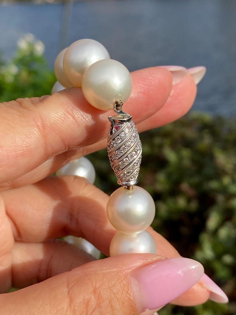 Pode incluir: Um close-up de um colar de p&eacute;rolas com um fecho decorativo. As p&eacute;rolas s&atilde;o grandes, redondas e brancas. O fecho &eacute; prateado e incrustado com pequenas pedras brilhantes. O fundo est&aacute; desfocado, com ind&iacute;cios de &aacute;gua e vegeta&ccedil;&atilde;o.