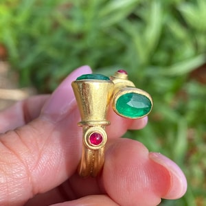 May include: A gold-toned bypass ring featuring two oval-cut green gemstones and two small, round, deep pink rubies set into the metal band. The ring has a matte, textured finish and an open design that allows the two ends to overlap slightly.