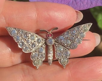 May include: A silver butterfly brooch with clear rhinestones and a red centre. The brooch is on a hand.