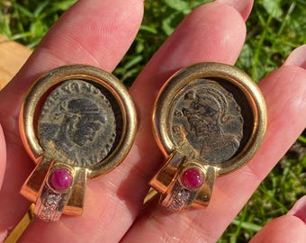 May include: A pair of gold earrings with a coin design and a ruby and diamond accent.