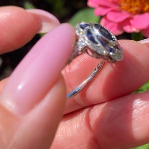 May include: A silver-toned ring featuring a central dark blue oval gemstone surrounded by a halo of small clear stones. The metal band is thin and polished, with intricate decorative metalwork visible along the sides of the setting.