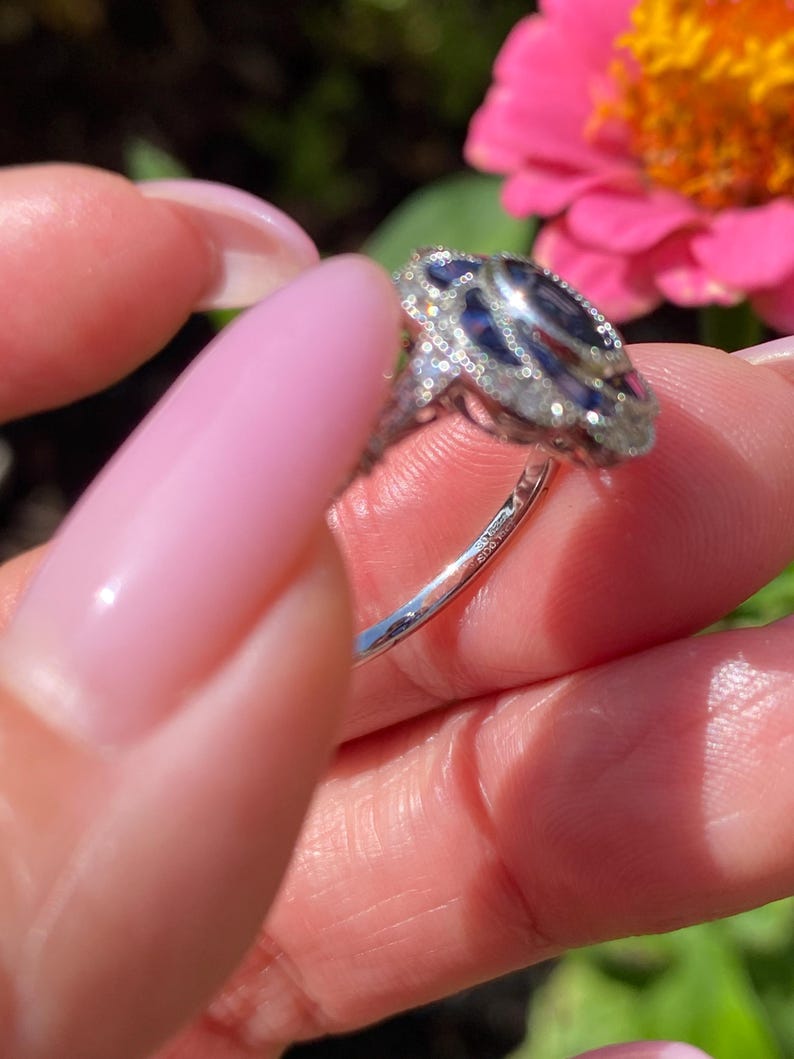 May include: A silver-toned ring featuring a central dark blue oval gemstone surrounded by a halo of small clear stones. The metal band is thin and polished, with intricate decorative metalwork visible along the sides of the setting.