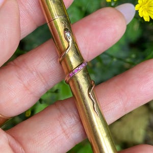 Op de afbeelding: Een cilindervormige goudkleurige metalen koker met een motief van een kronkelende slang om het lichaam. De slang heeft een kleine heldere steen in de kop en een band van kleine roze steentjes in het midden. Aan de bovenkant zit een metalen oogje.