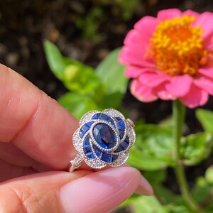 May include: A silver ring featuring a central oval-cut blue sapphire surrounded by a floral-shaped arrangement of smaller square-cut blue sapphires and tiny round white accent stones set in a metallic band with intricate milgrain detailing.