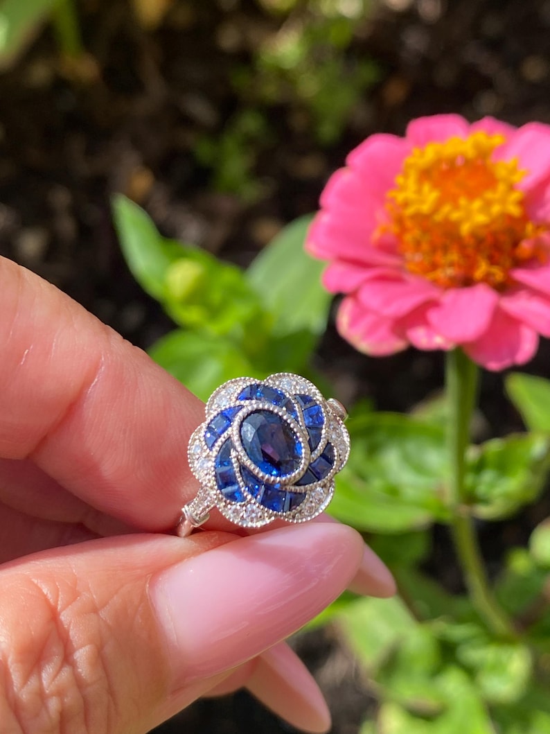 May include: A silver ring featuring a central oval-cut blue sapphire surrounded by a floral-shaped arrangement of smaller square-cut blue sapphires and tiny round white accent stones set in a metallic band with intricate milgrain detailing.