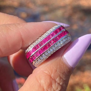 May include: A rectangular ring featuring three rows of vivid pink gemstones, framed by smaller white gemstones. The ring is held up against a blurred background, highlighting the ring's detailed design and bright colours.