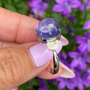 May include: A silver ring featuring a large, round, translucent, lavender-colored gemstone. The ring has a rectangular, light-colored stone beneath the main stone. The background is blurred with purple flowers and green foliage.