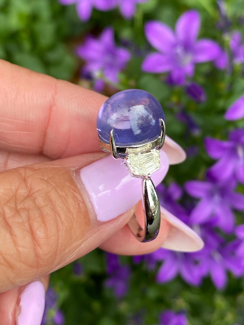 May include: A silver ring featuring a large, round, translucent, lavender-colored gemstone. The ring has a rectangular, light-colored stone beneath the main stone. The background is blurred with purple flowers and green foliage.