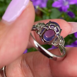 May include: A silver ring featuring a large, oval-shaped purple gemstone. The gemstone is surrounded by a decorative setting with smaller, rectangular purple stones. The ring is held up against a background of purple flowers.