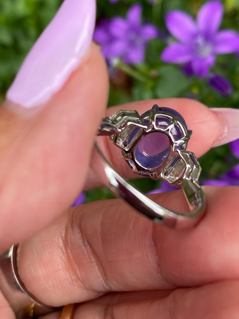 May include: A silver ring featuring a large, oval-shaped purple gemstone. The gemstone is surrounded by a decorative setting with smaller, rectangular purple stones. The ring is held up against a background of purple flowers.