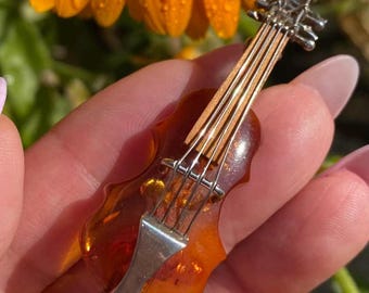 Op de afbeelding: Een miniatuur vioolbroche met een zilverkleurige metalen boog en snaren. De body van de viool is doorschijnend amberkleurig. De broche wordt in een hand gehouden, met een vage achtergrond van een oranje bloem.