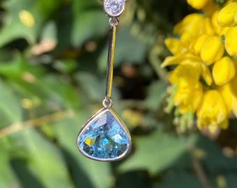 Op de afbeelding: Een zilveren ketting met een druppelvormige, lichtblauwe edelsteen hanger. De hanger hangt aan een dunne, verticale staaf en een kleine, ronde, heldere edelsteen. De achtergrond toont gele bloemen en groen gebladerte.