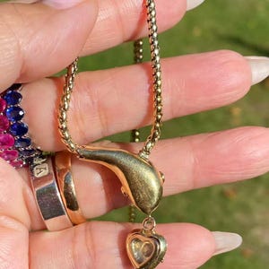 May include: A gold-toned necklace with a snake-shaped pendant and a heart-shaped charm. The necklace has a delicate chain and is displayed against a blurred green background. Other gold rings and a ring with blue and pink stones are also visible.