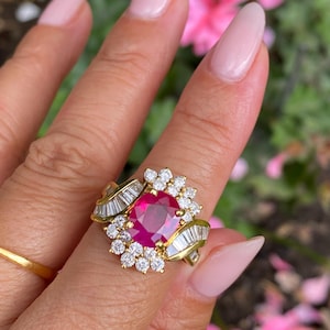 Może przedstawiać: Złoty pierścionek z dużym, żywym czerwonym kamieniem szlachetnym pośrodku, otoczonym mniejszymi białymi kamieniami szlachetnymi i diamentami w kształcie bagietki. Pierścionek ma szczegółowy, ozdobny wzór.