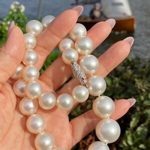 Pode incluir: Um colar de p&eacute;rolas brancas grandes e brilhantes. As p&eacute;rolas s&atilde;o enfiadas e presas com um fecho prateado adornado com pequenas pedras brilhantes. O colar &eacute; segurado contra um fundo desfocado ao ar livre.