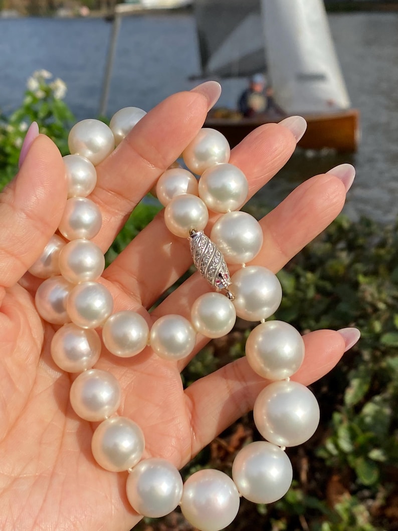 Pode incluir: Um colar de p&eacute;rolas brancas grandes e brilhantes. As p&eacute;rolas s&atilde;o enfiadas e presas com um fecho prateado adornado com pequenas pedras brilhantes. O colar &eacute; segurado contra um fundo desfocado ao ar livre.