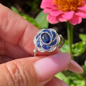 May include: A silver-toned ring featuring a central oval-cut blue sapphire surrounded by a floral-shaped halo of smaller baguette-cut blue sapphires and tiny clear round stones, set in a scalloped metal frame with a polished silver band.