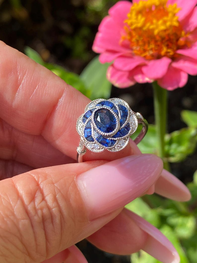 May include: A silver-toned ring featuring a central oval-cut blue sapphire surrounded by a floral-shaped halo of smaller baguette-cut blue sapphires and tiny clear round stones, set in a scalloped metal frame with a polished silver band.