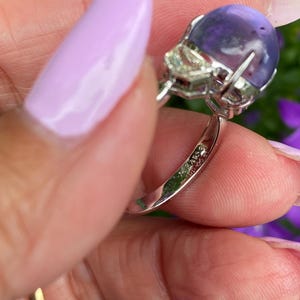 May include: A close-up of a silver ring featuring a large, round, purple gemstone. The ring also has a smaller, clear, square-cut stone. The ring is held by a person with light purple nail polish.