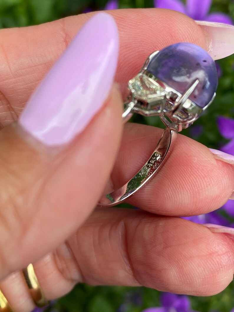 May include: A close-up of a silver ring featuring a large, round, purple gemstone. The ring also has a smaller, clear, square-cut stone. The ring is held by a person with light purple nail polish.