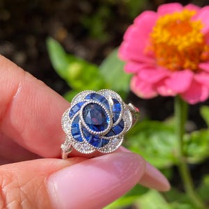 May include: A silver ring featuring a central oval-cut blue sapphire surrounded by a floral-shaped halo of smaller square-cut blue sapphires and tiny round white accent stones, all set in a detailed metal frame with a milgrain edge finish.