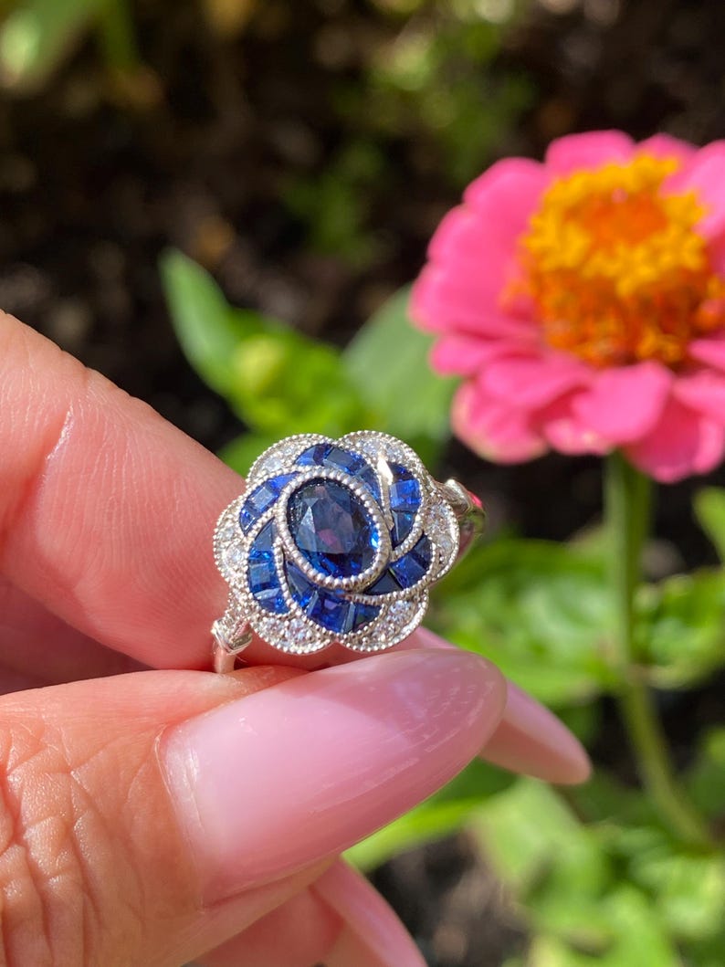 May include: A silver ring featuring a central oval-cut blue sapphire surrounded by a floral-shaped halo of smaller square-cut blue sapphires and tiny round white accent stones, all set in a detailed metal frame with a milgrain edge finish.