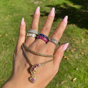 May include: A hand displays various pieces of jewelry against a green background. Rings with diamonds and sapphires adorn the fingers, while a gold necklace with a ruby pendant rests in the palm. The jewelry sparkles in the sunlight.