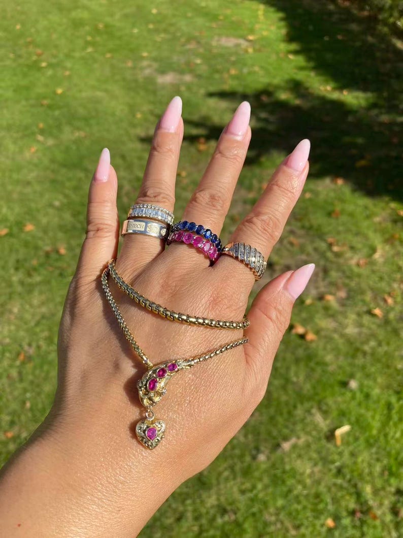 May include: A hand displays various pieces of jewelry against a green background. Rings with diamonds and sapphires adorn the fingers, while a gold necklace with a ruby pendant rests in the palm. The jewelry sparkles in the sunlight.