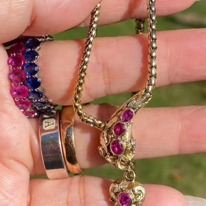 May include: A gold necklace with a pendant featuring a heart and three round gemstones. The necklace is held up by a hand, and there are several rings on the fingers, including a ring with pink and blue gemstones.