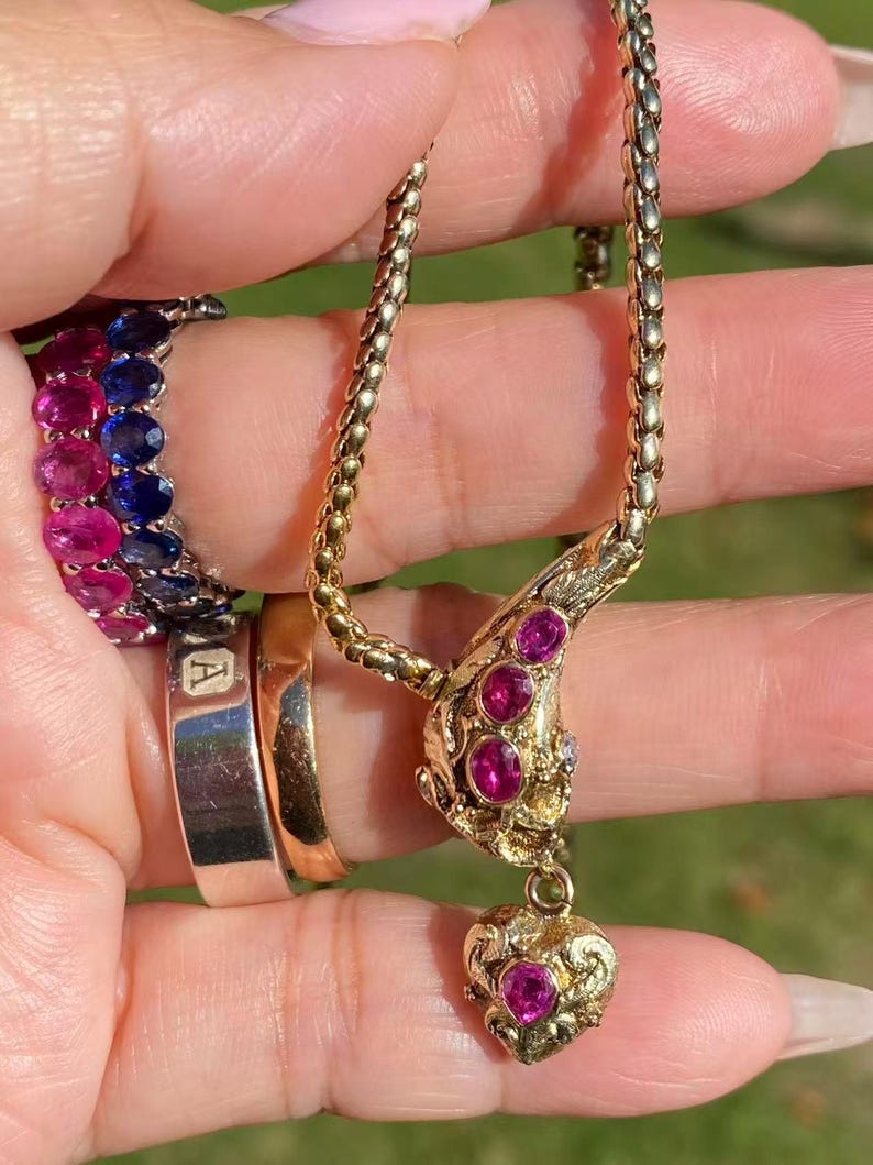 May include: A gold necklace with a pendant featuring a heart and three round gemstones. The necklace is held up by a hand, and there are several rings on the fingers, including a ring with pink and blue gemstones.