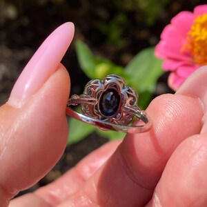 May include: A silver-toned ring featuring an oval-shaped dark blue gemstone set in a decorative, openwork metal frame. The setting has a scalloped edge design surrounding the central stone, with a smooth, polished band extending from the sides.