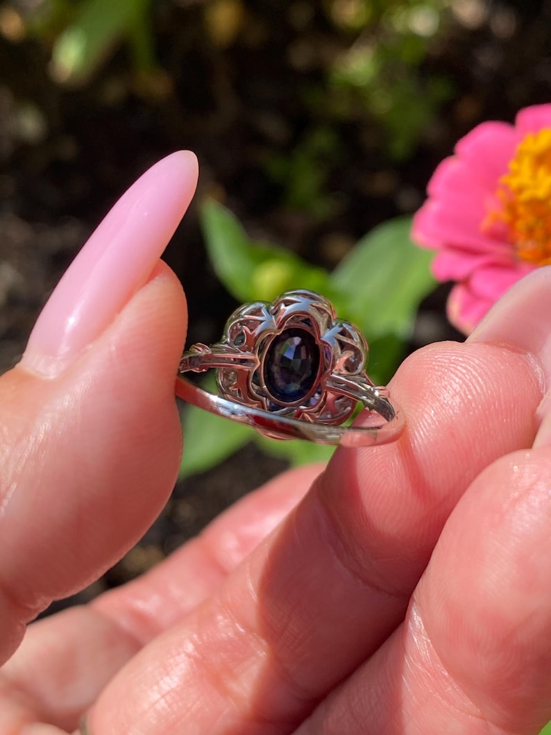 May include: A silver-toned ring featuring an oval-shaped dark blue gemstone set in a decorative, openwork metal frame. The setting has a scalloped edge design surrounding the central stone, with a smooth, polished band extending from the sides.