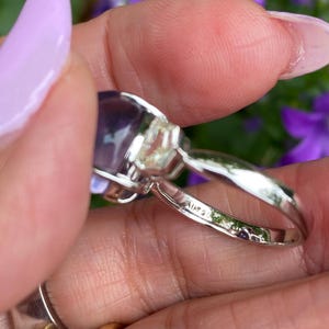 May include: A silver ring with a large, oval, purple gemstone. The ring has a delicate band and is held up by a hand with a light purple nail polish. The ring is set against a background of purple flowers.