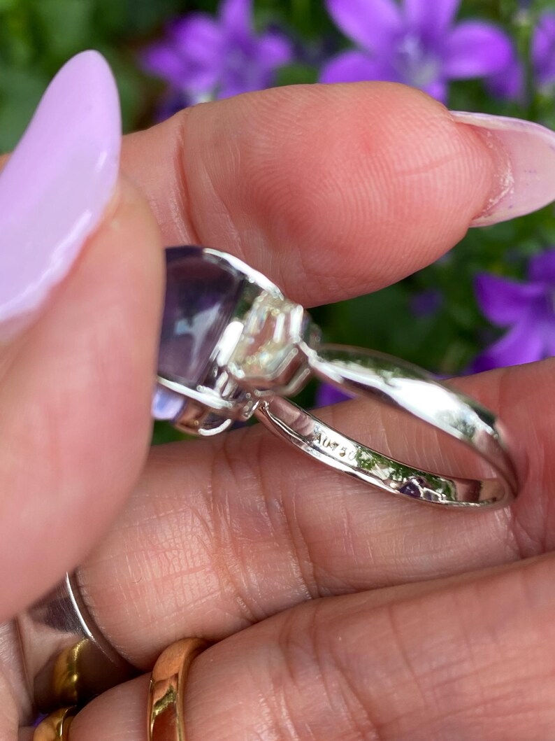 May include: A silver ring with a large, oval, purple gemstone. The ring has a delicate band and is held up by a hand with a light purple nail polish. The ring is set against a background of purple flowers.