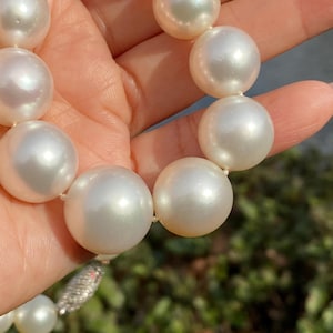 Pode incluir: Um colar de p&eacute;rolas brancas grandes e lustrosas. As p&eacute;rolas s&atilde;o redondas e de tamanho uniforme, enfiadas com um fio delicado. O fecho &eacute; prateado com pequenos detalhes brilhantes. As p&eacute;rolas est&atilde;o sendo seguradas em uma m&atilde;o.