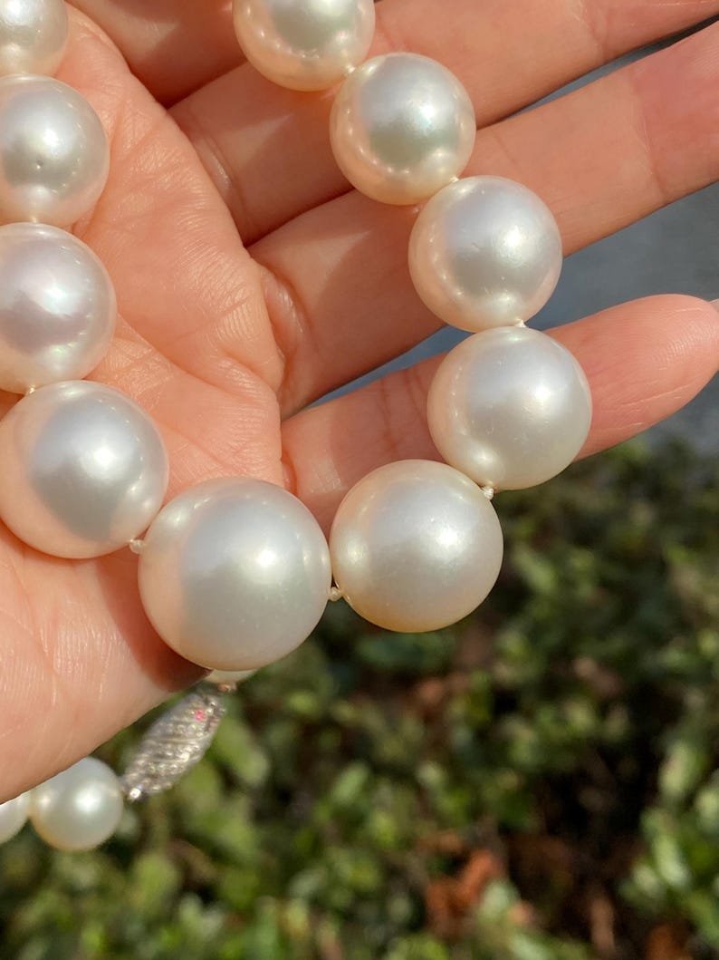 Pode incluir: Um colar de p&eacute;rolas brancas grandes e lustrosas. As p&eacute;rolas s&atilde;o redondas e de tamanho uniforme, enfiadas com um fio delicado. O fecho &eacute; prateado com pequenos detalhes brilhantes. As p&eacute;rolas est&atilde;o sendo seguradas em uma m&atilde;o.