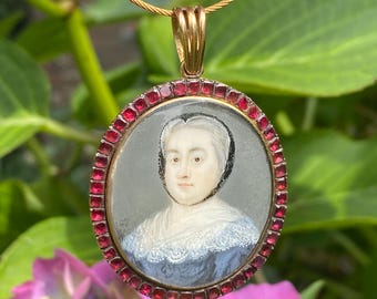 May include: A gold pendant necklace with a portrait miniature of a woman in a white lace collar and a blue dress. The miniature is surrounded by a border of red gemstones.