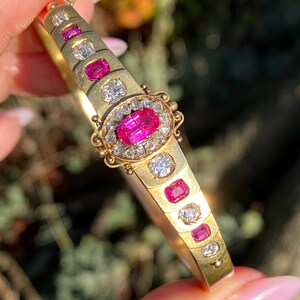 May include: A gold bracelet with a central oval-shaped pink gemstone surrounded by smaller white gemstones. The bracelet features alternating pink and white gemstones along its length, set in a gold band. The bracelet is held up against a blurred green background.