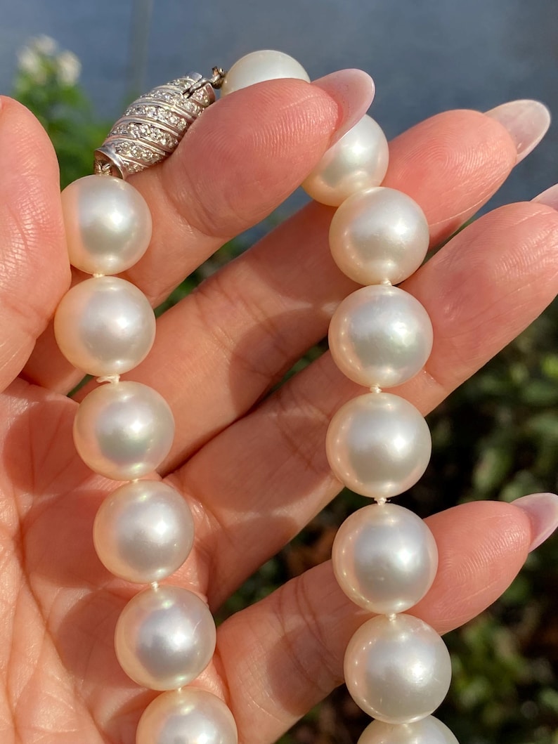 Pode incluir: Close-up de um colar de p&eacute;rolas. O colar apresenta grandes p&eacute;rolas brancas redondas e brilhantes, enfiadas juntas. O fecho &eacute; prateado e adornado com pequenas pedras brilhantes. As p&eacute;rolas s&atilde;o seguradas em uma m&atilde;o.