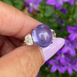 May include: A silver ring featuring a large, oval, lavender-colored gemstone as the centerpiece. Two rectangular, clear gemstones flank the main stone. The ring is held against a backdrop of purple flowers.