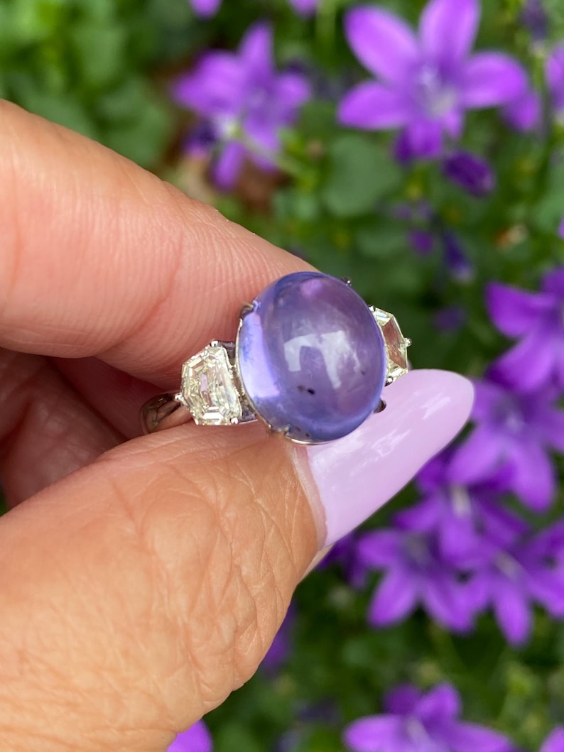 May include: A silver ring featuring a large, oval, lavender-colored gemstone as the centerpiece. Two rectangular, clear gemstones flank the main stone. The ring is held against a backdrop of purple flowers.