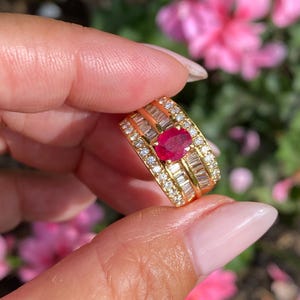 May include: A gold ring with a central oval ruby, encircled by smaller diamonds and baguette-cut stones. The ring has a layered design with a polished finish. The ring is held up against a backdrop of pink flowers, with a bright, natural light.